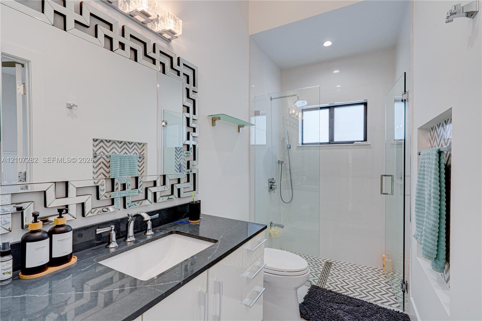Glossy white cabinetry, dark stone countertop with an undermount sink, geometric mirror frame, walk-in glass-enclosed shower with chevron-patterned tile flooring, and integrated shower wall niche