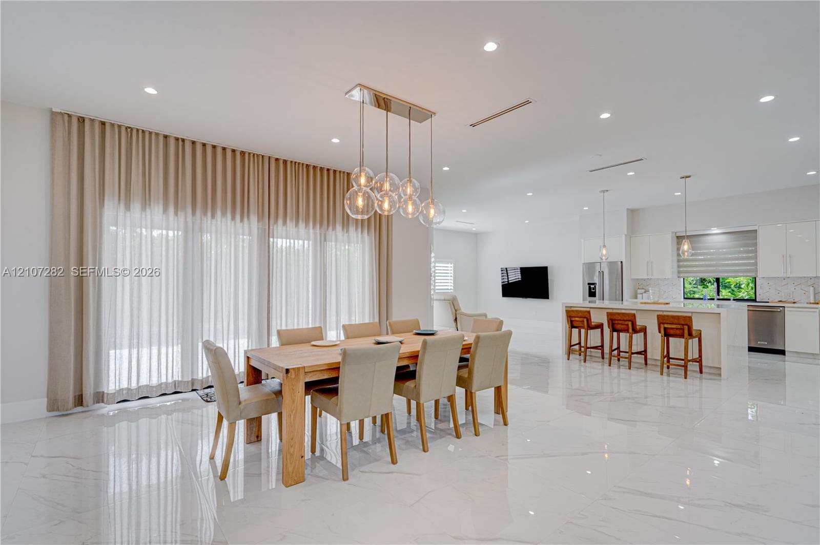 Open-concept interior featuring large-format polished stone flooring, recessed ceiling lighting, a multi-globe linear chandelier, and a kitchen island with integrated stainless steel appliances