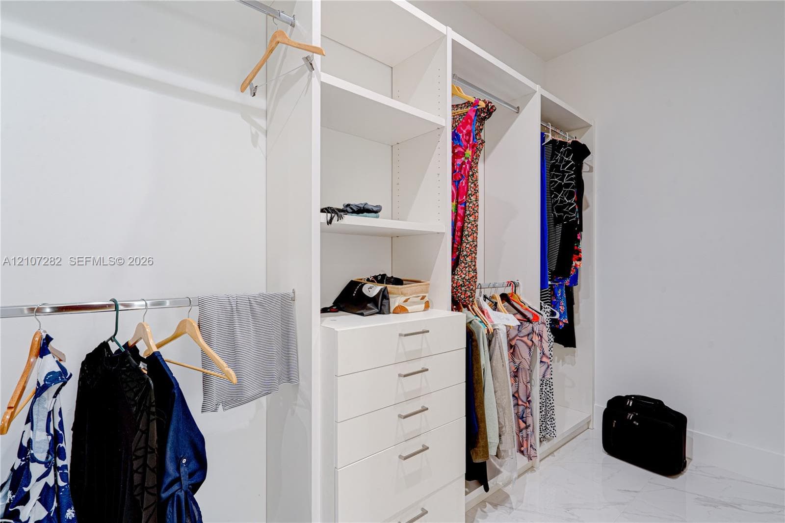 Custom walk-in closet featuring white built-in shelving, a central column of five soft-close drawers, stainless steel hanging rods, and polished stone-finish flooring