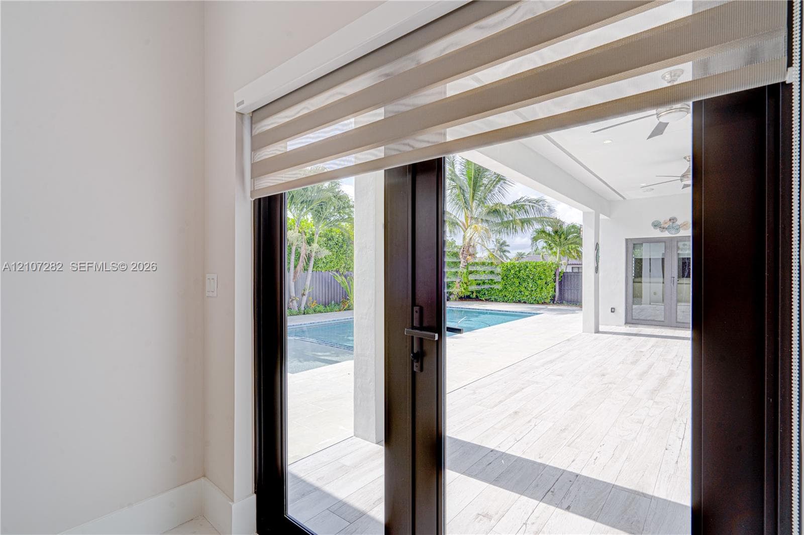 Dark-framed glass double doors featuring dual-layer roller shades, opening onto a covered patio with light-toned tile flooring, recessed ceiling lighting, and a swimming pool