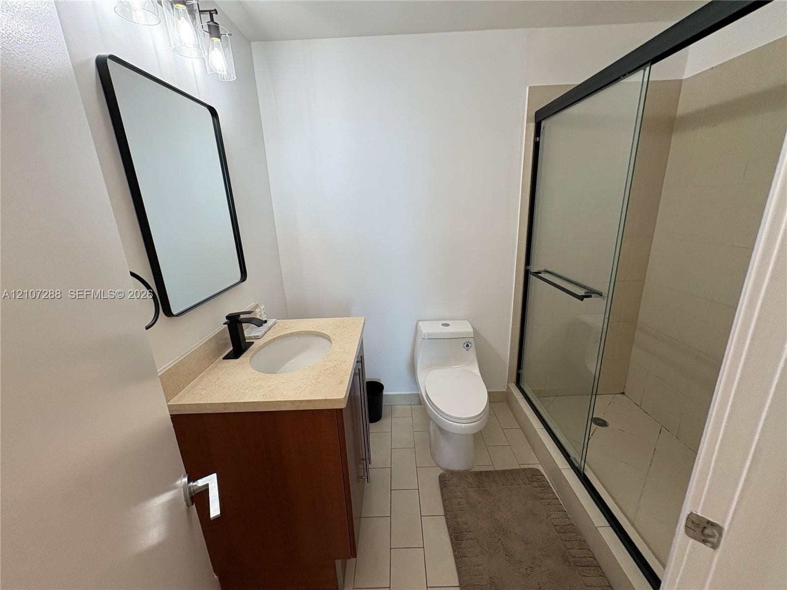 Wood-finish vanity with a light-toned countertop, undermount sink, matte black faucet, rectangular mirror with a black frame, and a walk-in shower featuring a sliding glass enclosure