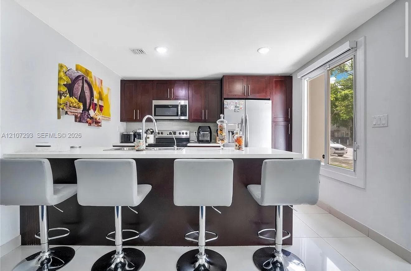 Kitchen featuring dark wood cabinetry, white countertops, stainless steel appliances, a center island with a stainless steel faucet, and tile flooring