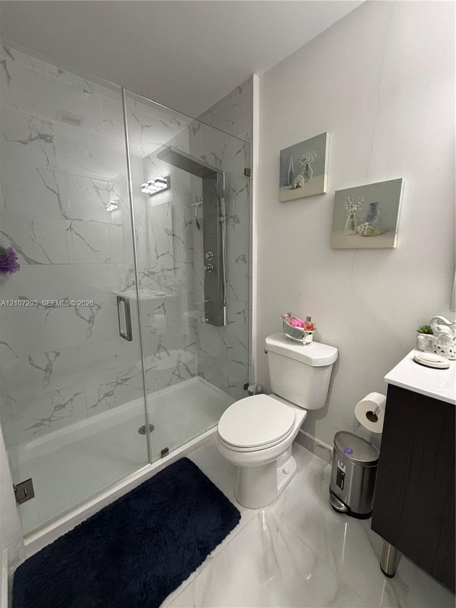 Walk-in shower featuring glass paneling, floor-to-ceiling stone-style wall tile, and an integrated multi-jet shower panel, complemented by a white vanity with a solid-surface countertop and large-format porcelain tile flooring