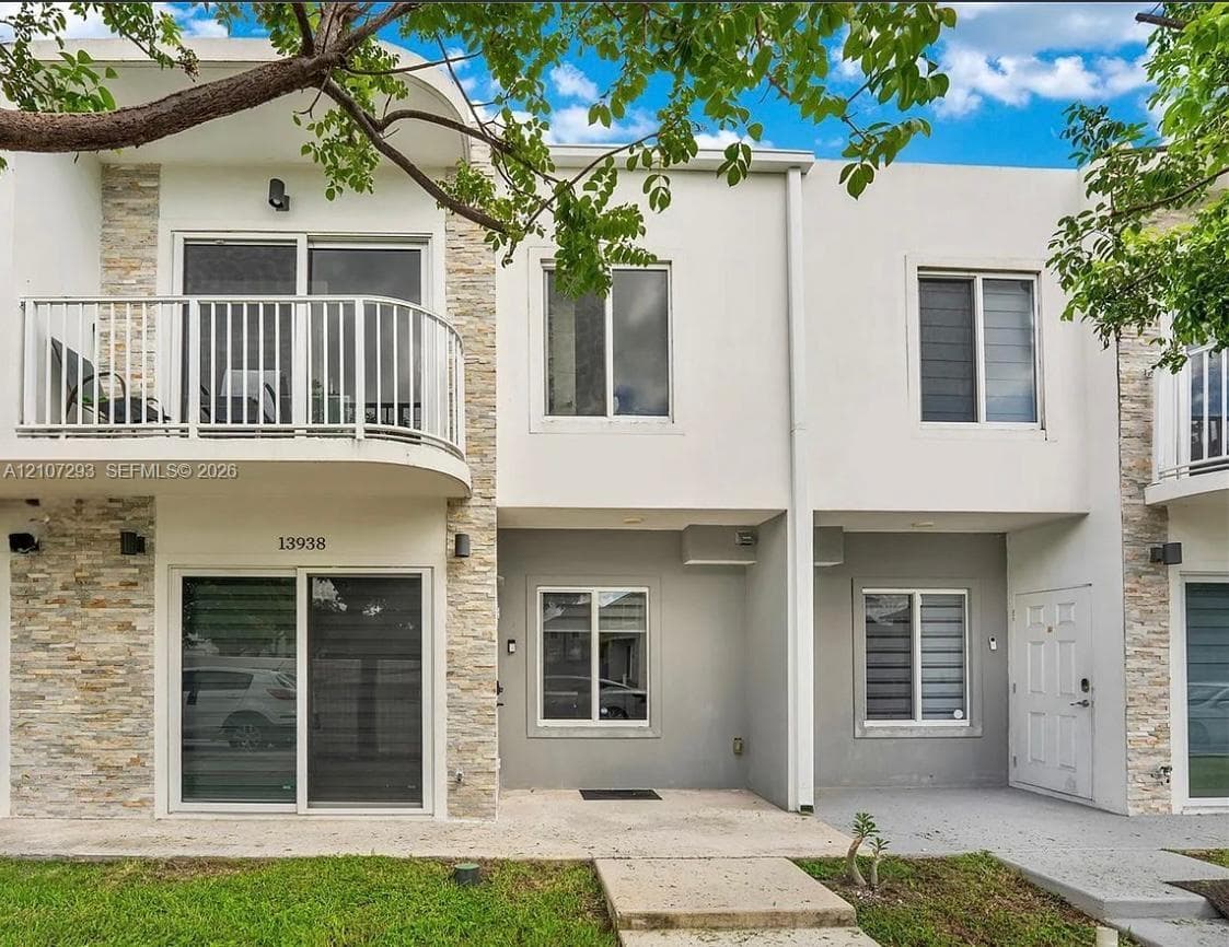 Modern townhome exterior featuring stacked stone accents, second-story balcony with white metal railing, dual-pane sliding glass doors, and recessed exterior lighting
