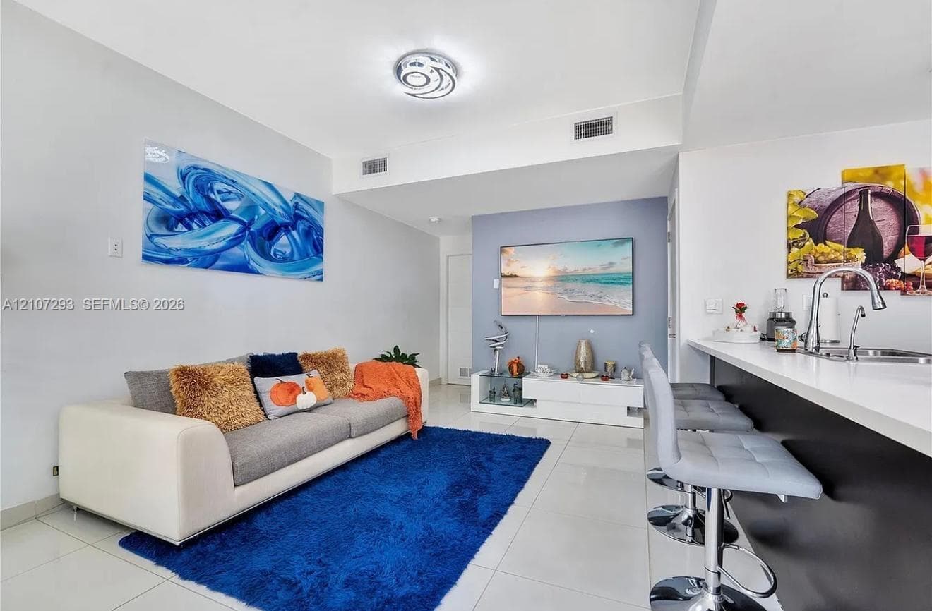 Neutral-toned living space featuring large-format tile flooring, accent wall, modern ceiling-mounted light fixture, and kitchen breakfast bar with countertop sink