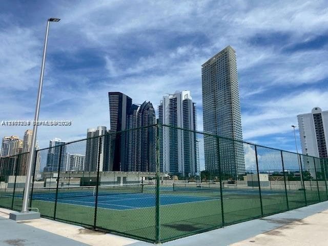 Hard-surfaced tennis court enclosed by chain-link fencing, featuring overhead pole lighting