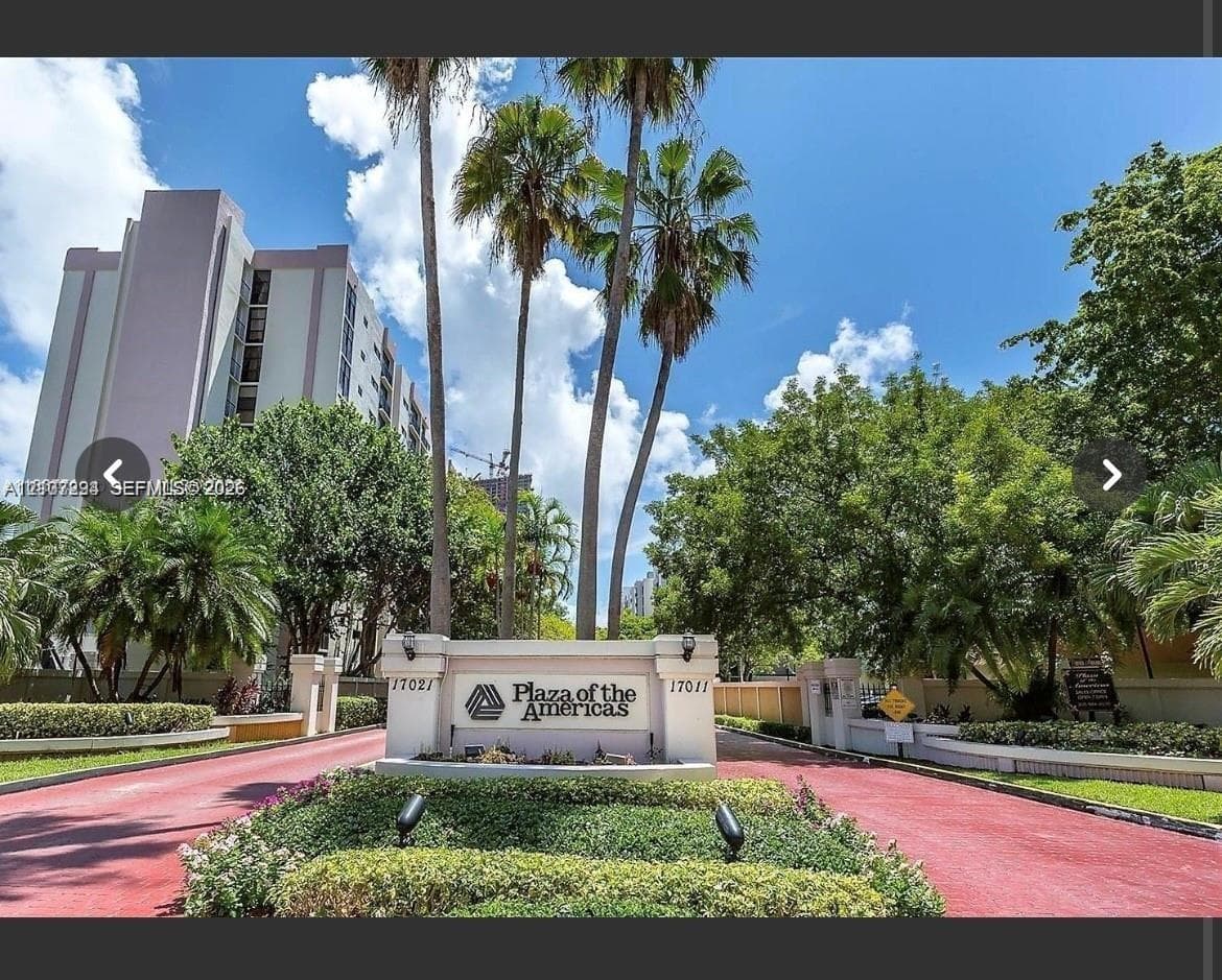 Gated entrance monument with integrated lighting, paved driveway, mature palm trees, and professional landscaping