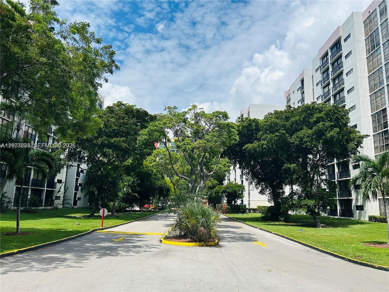 Multi-story residential complex featuring white stucco facades, private balconies, expansive green space, mature canopy trees, and paved thoroughfares