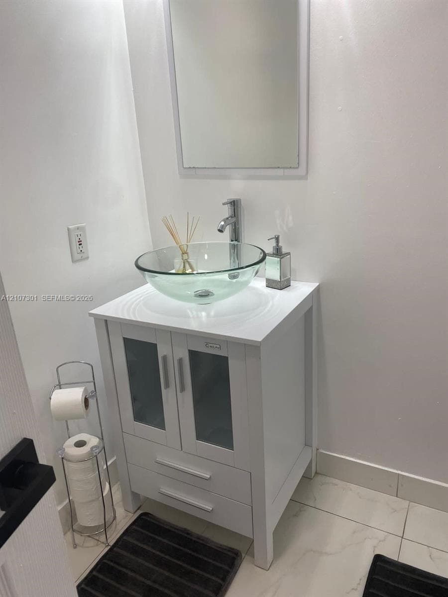 White vanity featuring cabinet storage with glass panel doors, a circular clear glass vessel sink, chrome single-handle faucet, and large-format tile flooring