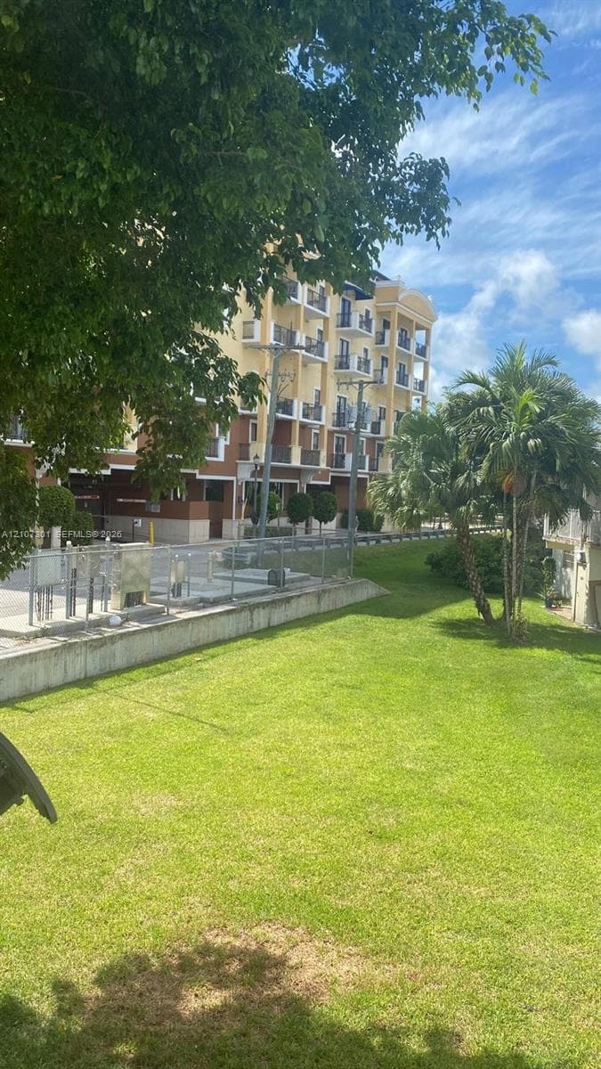 Multi-story exterior building featuring balconies with railings, concrete foundation, metal fencing, and landscaped grounds with mature trees