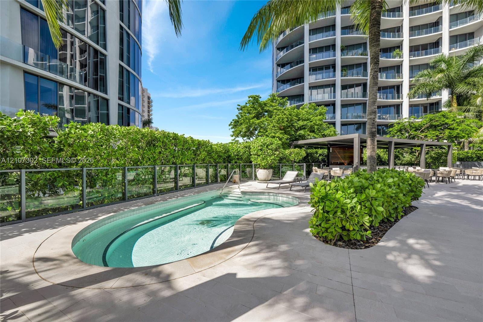 Curved swimming pool featuring a glass pane perimeter fence, stone-finish decking, and a covered pavilion structure