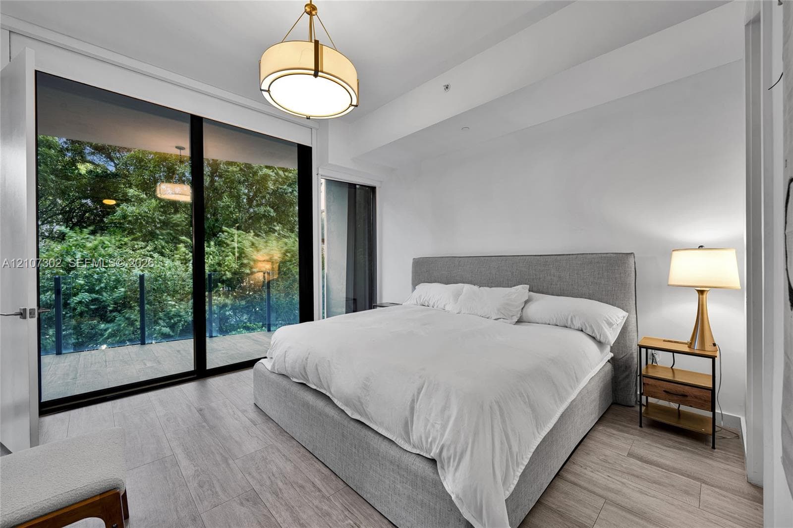 Floor-to-ceiling glass sliding doors opening to an exterior balcony, light-toned wood-finish flooring, and a circular drum pendant light fixture