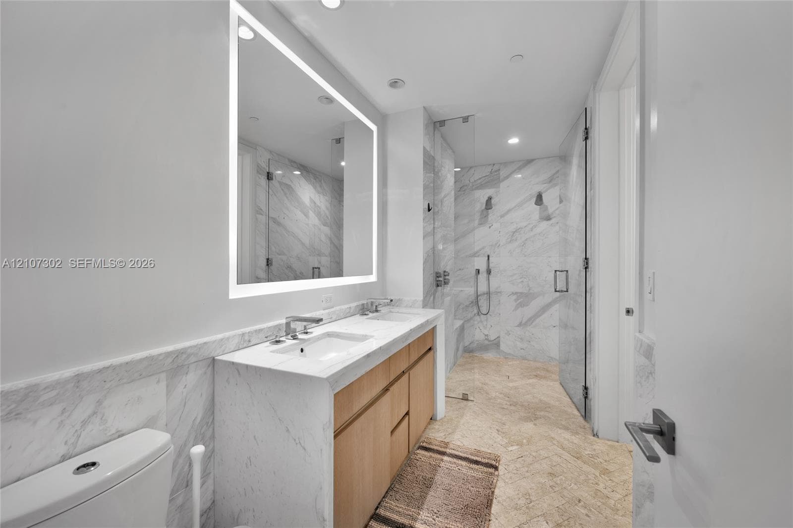 Dual-vanity cabinetry with stone countertop, integrated light-up mirror, walk-in shower with stone wall tile, and herringbone-patterned stone flooring