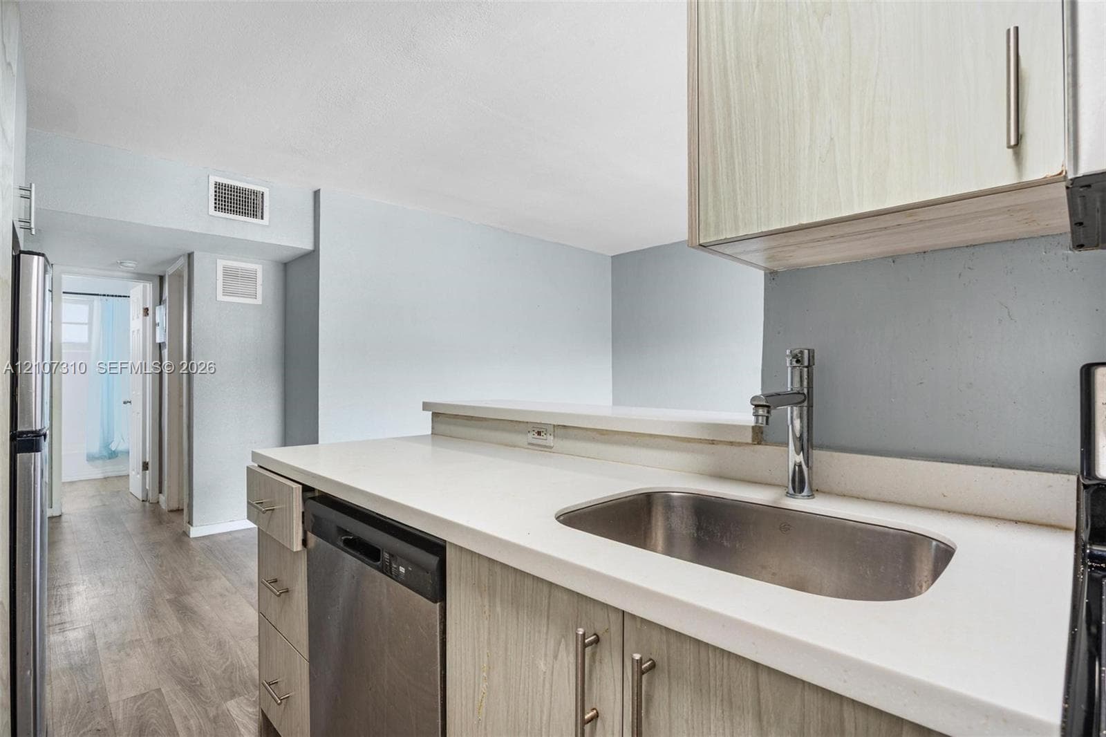 Kitchen featuring light-tone wood-finish cabinetry, white solid-surface countertops, stainless steel undermount sink with high-arc faucet, stainless steel dishwasher, and wood-finish flooring