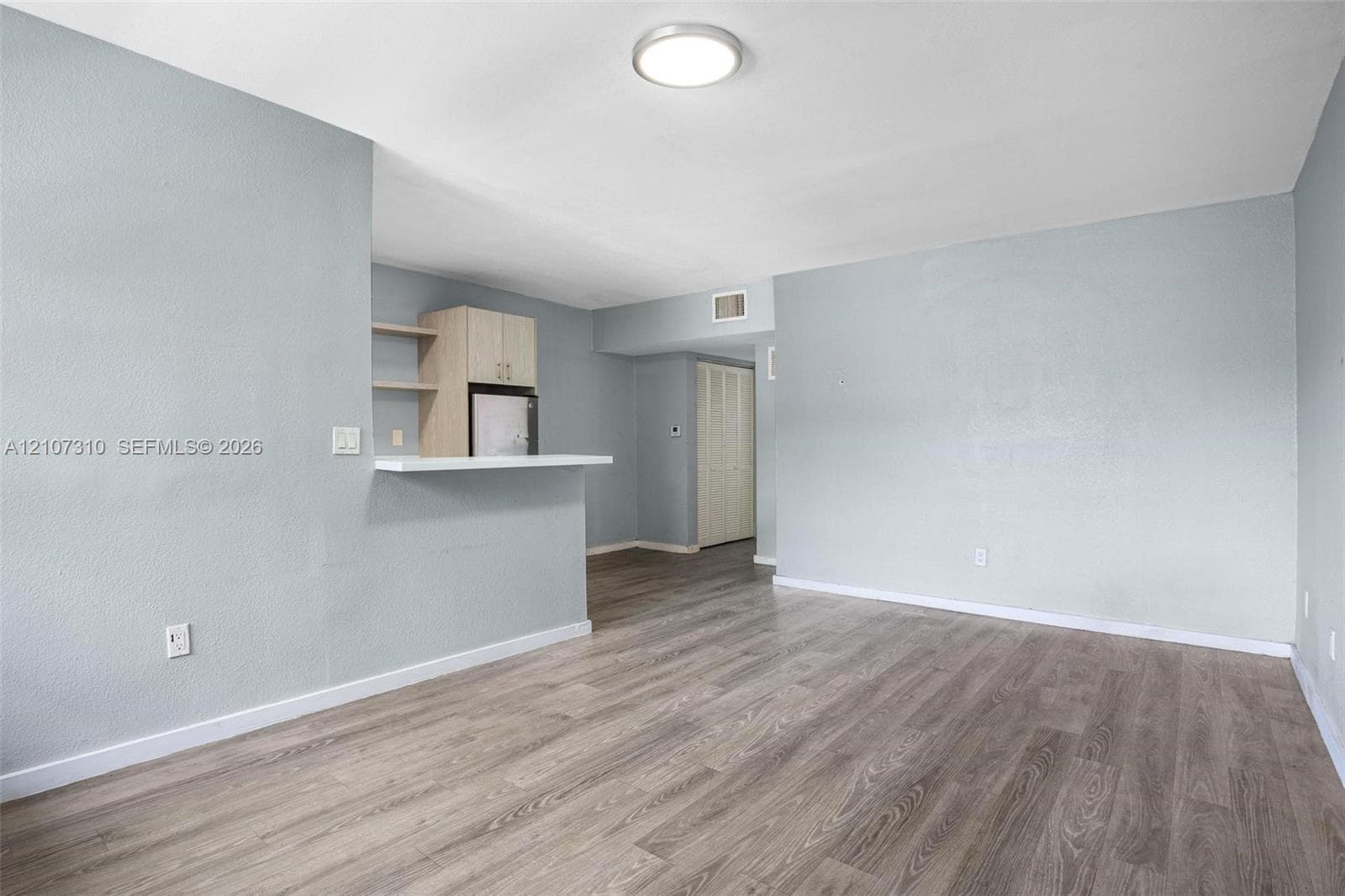 Neutral-toned living space featuring wood-finish flooring, a pass-through breakfast bar, built-in upper cabinetry, and a flush-mount overhead light fixture