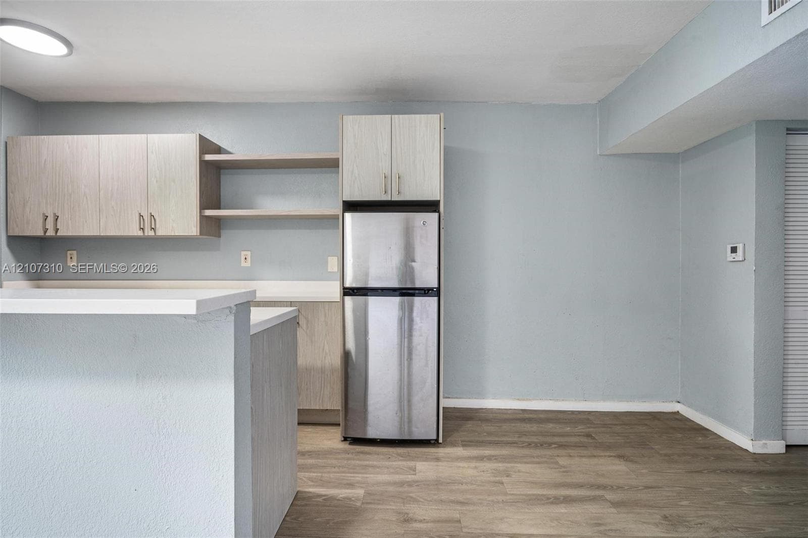 Kitchen area featuring wood-finish cabinetry, open shelving, a white countertop, a stainless steel refrigerator, and wood-finish flooring