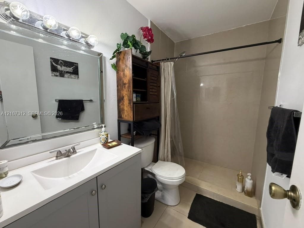 Bathroom featuring a vanity with a white countertop and integrated sink, four-bulb vanity lighting fixture, wall-mounted mirror, and a walk-in shower with tiled walls and flooring