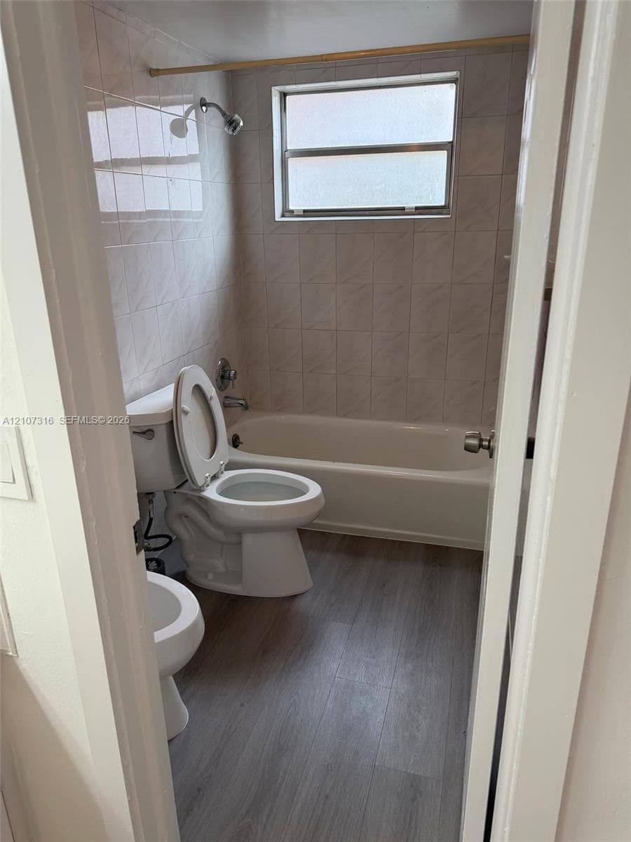 Bathroom featuring wood-finish flooring, a white bathtub with a tile surround, and a horizontal window