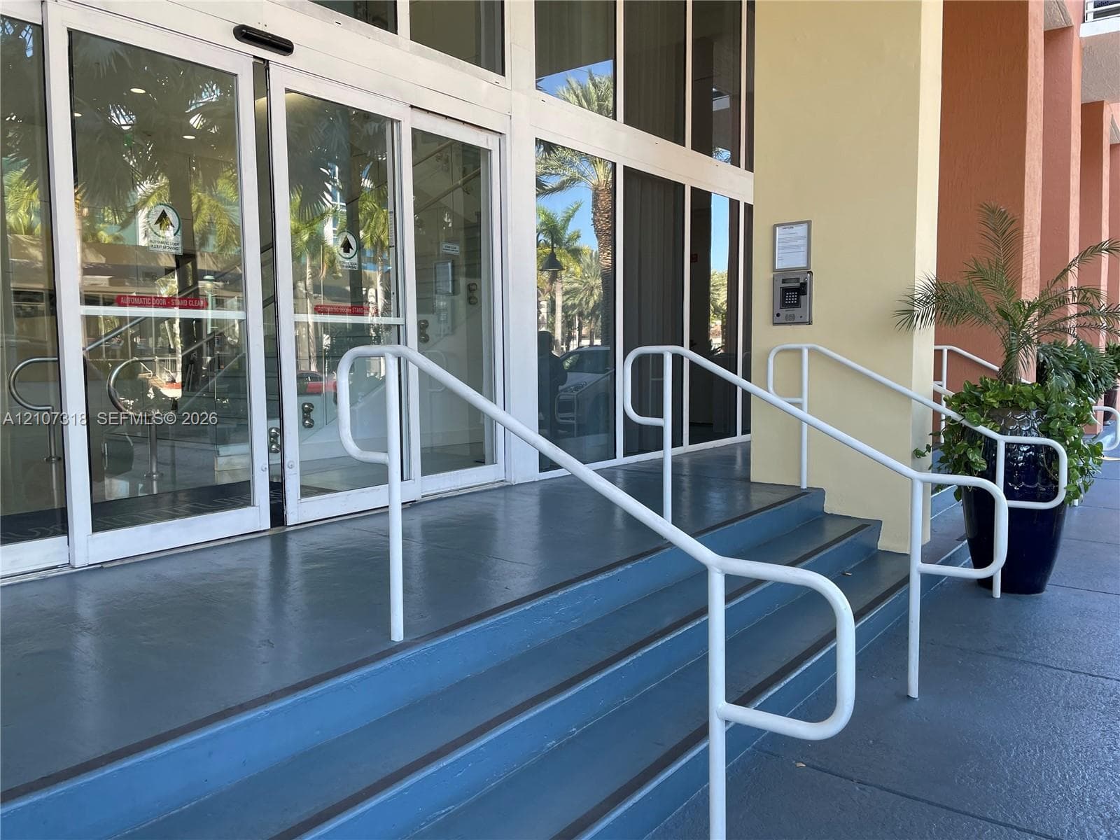 Building entrance featuring automatic glass sliding doors, structural white metal railings, and a secure electronic entry system