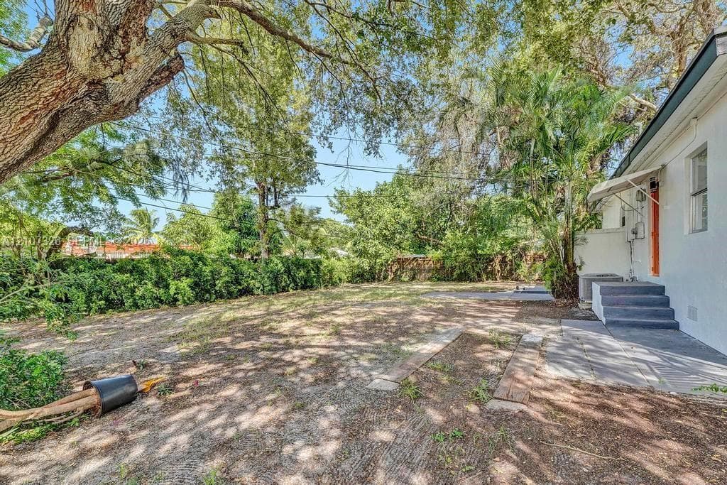 Backyard space featuring mature shade trees, established perimeter shrubbery, concrete patio area, and elevated concrete steps with a metal awning
