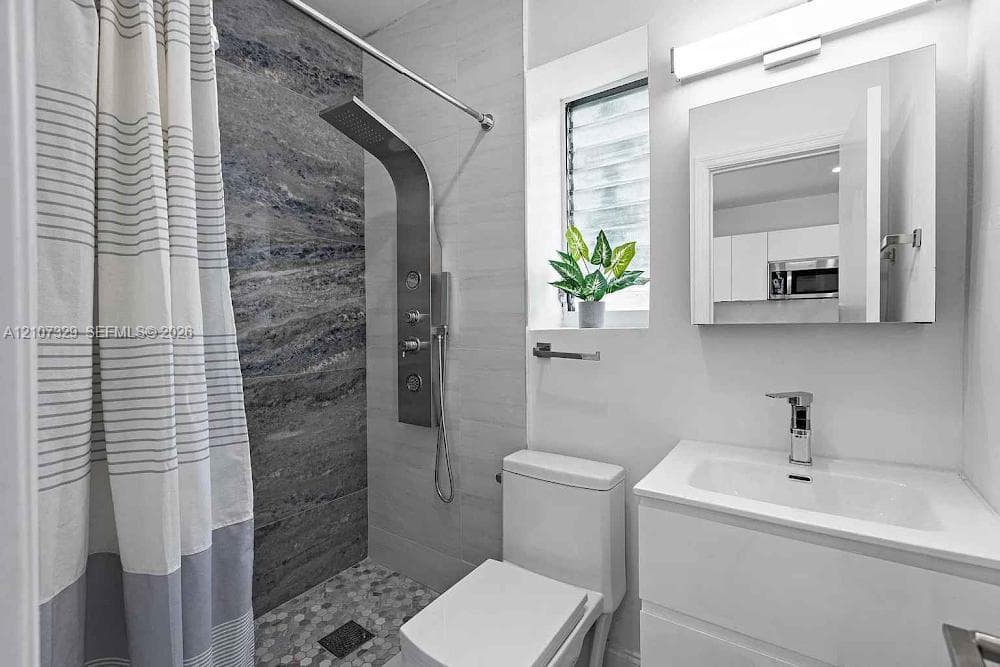 Shower stall featuring a multi-function stainless steel panel system and textured stone-finish tile surround with pebble-style floor tiling, complemented by a modern white vanity with integrated sink and mirrored medicine cabinet