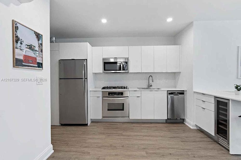 Kitchen featuring white flat-panel cabinetry, stainless steel appliances, a gas cooktop, quartz countertops, and an under-counter beverage refrigerator, set over wood-finish flooring