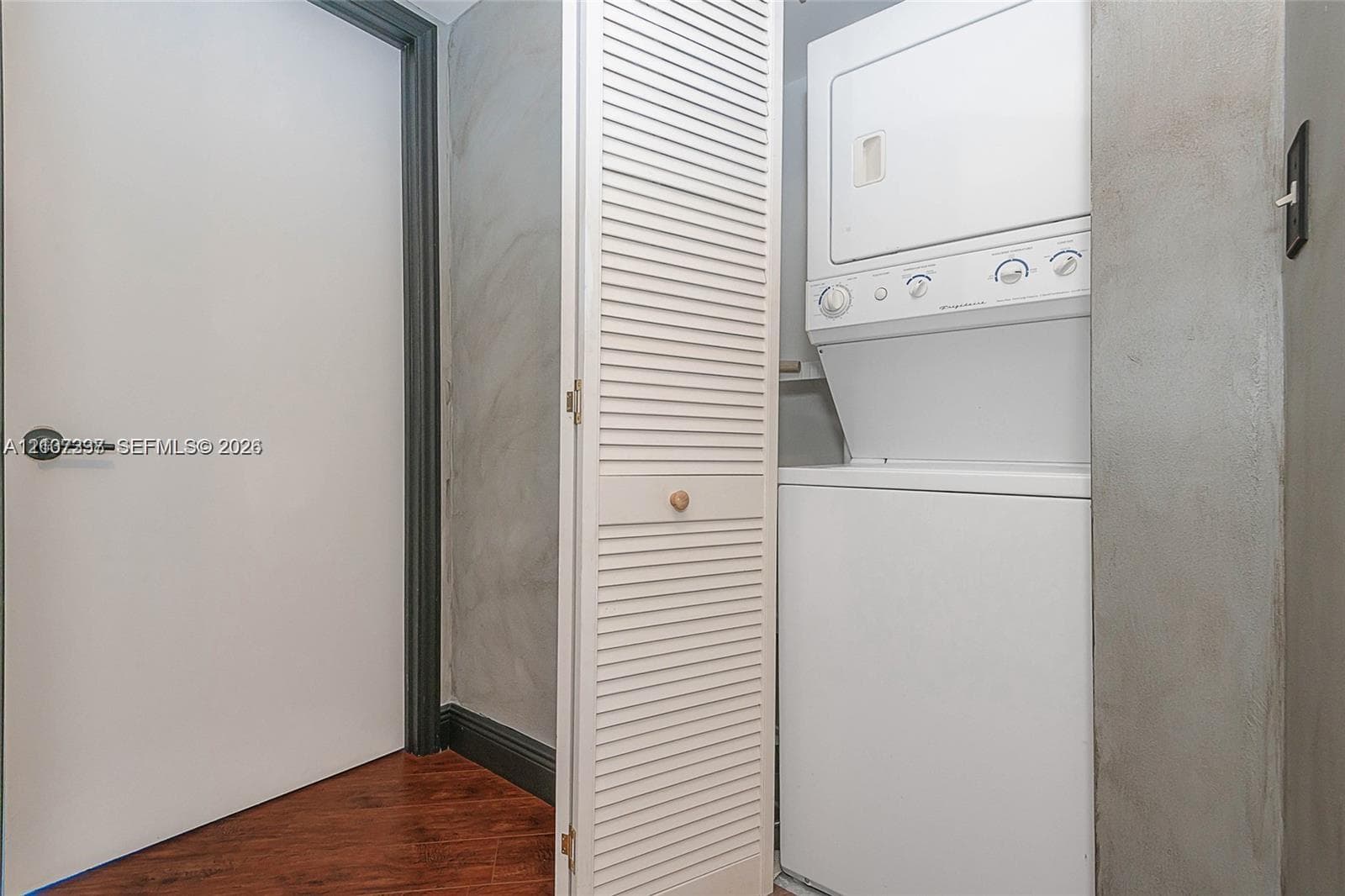 Laundry closet featuring a white stacked washer and dryer unit, louvered bi-fold door, dark trim accents, and wood-finish flooring