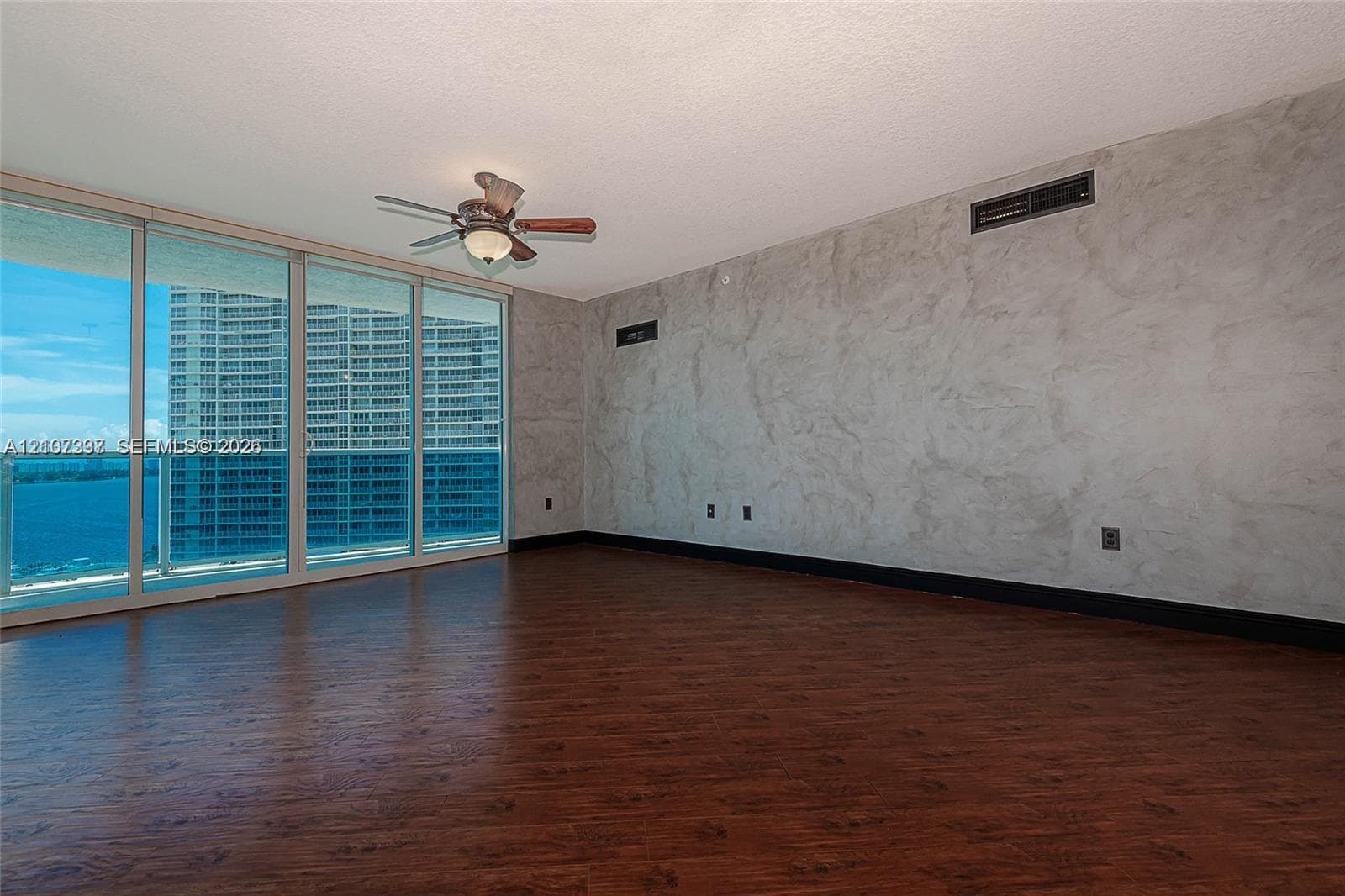 Floor-to-ceiling sliding glass doors, dark wood-finish flooring, decorative plaster wall finish, and a ceiling-mounted lighting fixture with an integrated fan