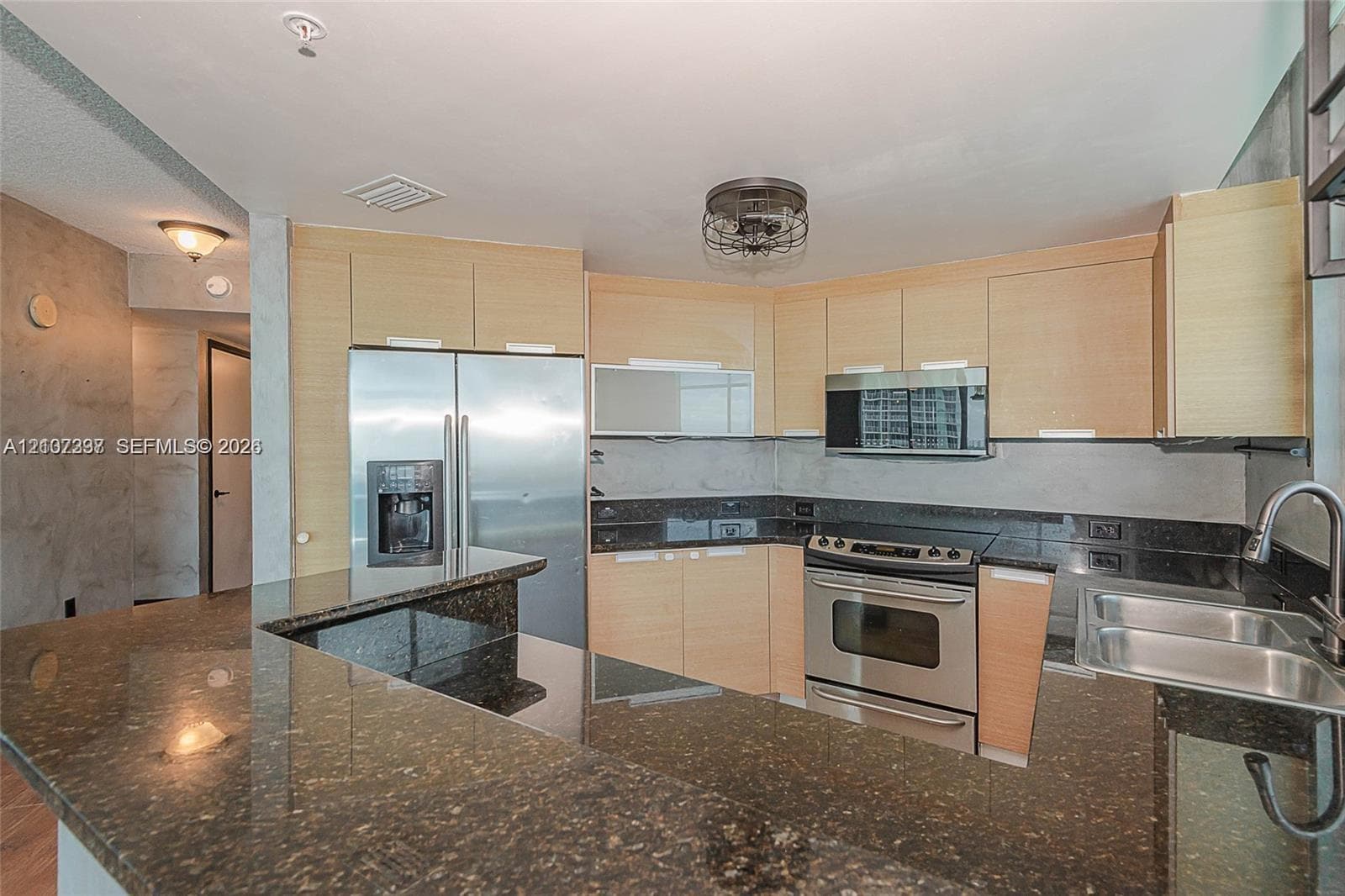 Kitchen featuring flat-panel light wood cabinetry, dark stone countertops, stainless steel appliances, breakfast bar, and stainless steel dual-basin sink