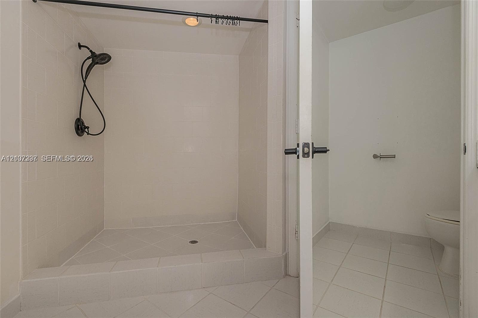 Walk-in shower featuring white tiled walls, a matte black handheld showerhead, a raised threshold, and white floor tile throughout