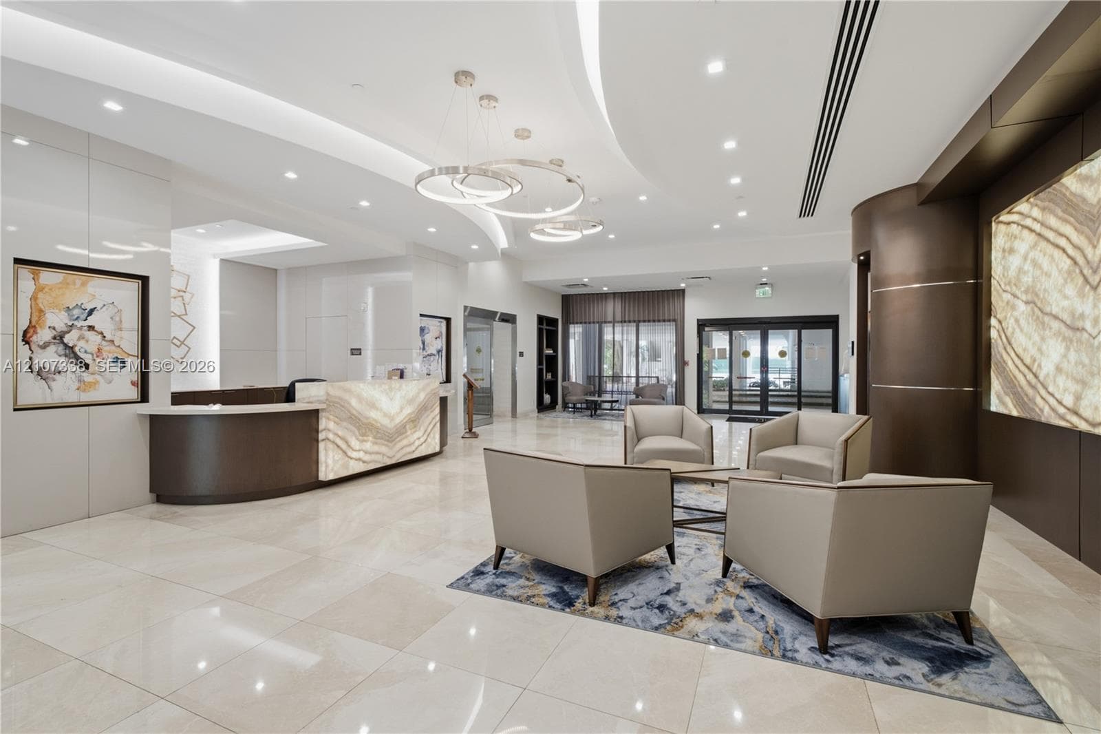 High-ceiling lobby featuring a backlit stone-front reception desk, polished stone flooring, recessed lighting, and contemporary ring chandeliers