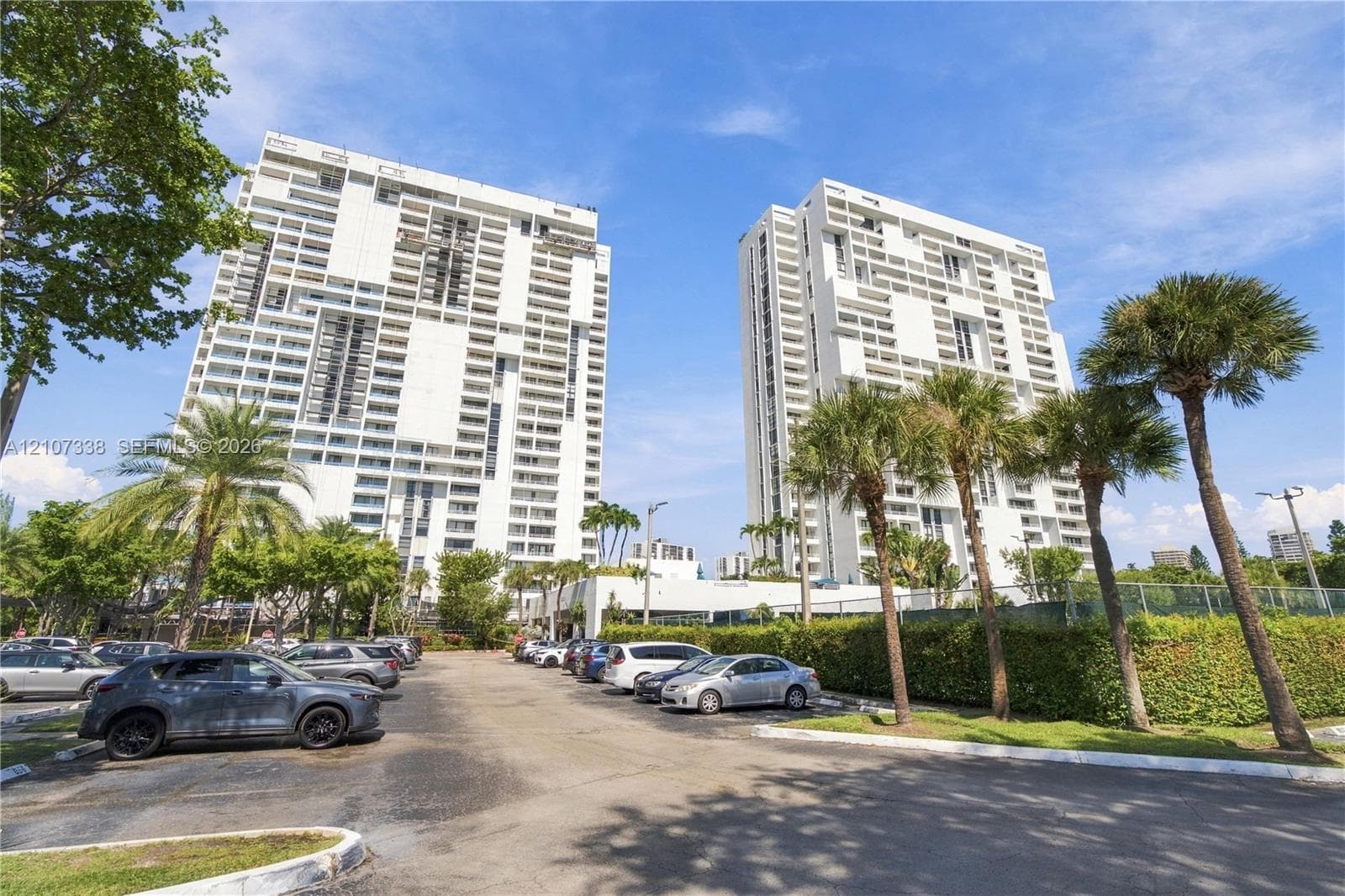 High-rise residential towers featuring white concrete facades, multiple levels of private balconies, and an extensive parking area with mature palm trees