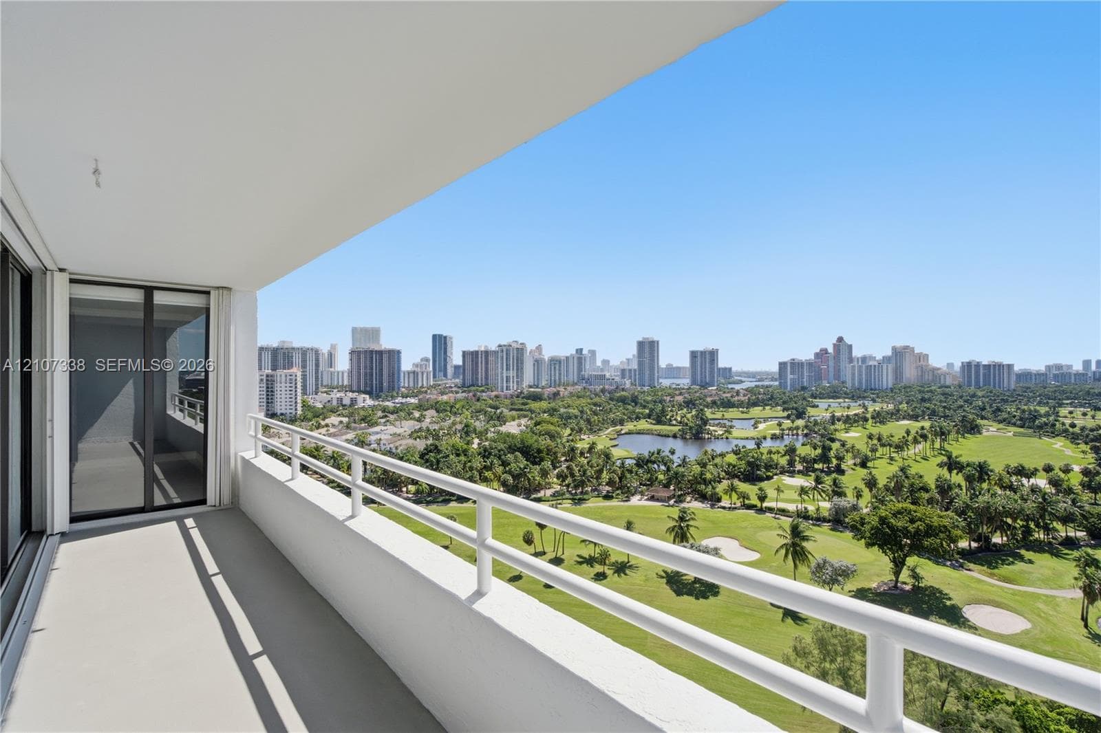 Covered balcony featuring white railings, neutral flooring, and glass sliding door access, showcasing expansive golf course and city skyline scenery