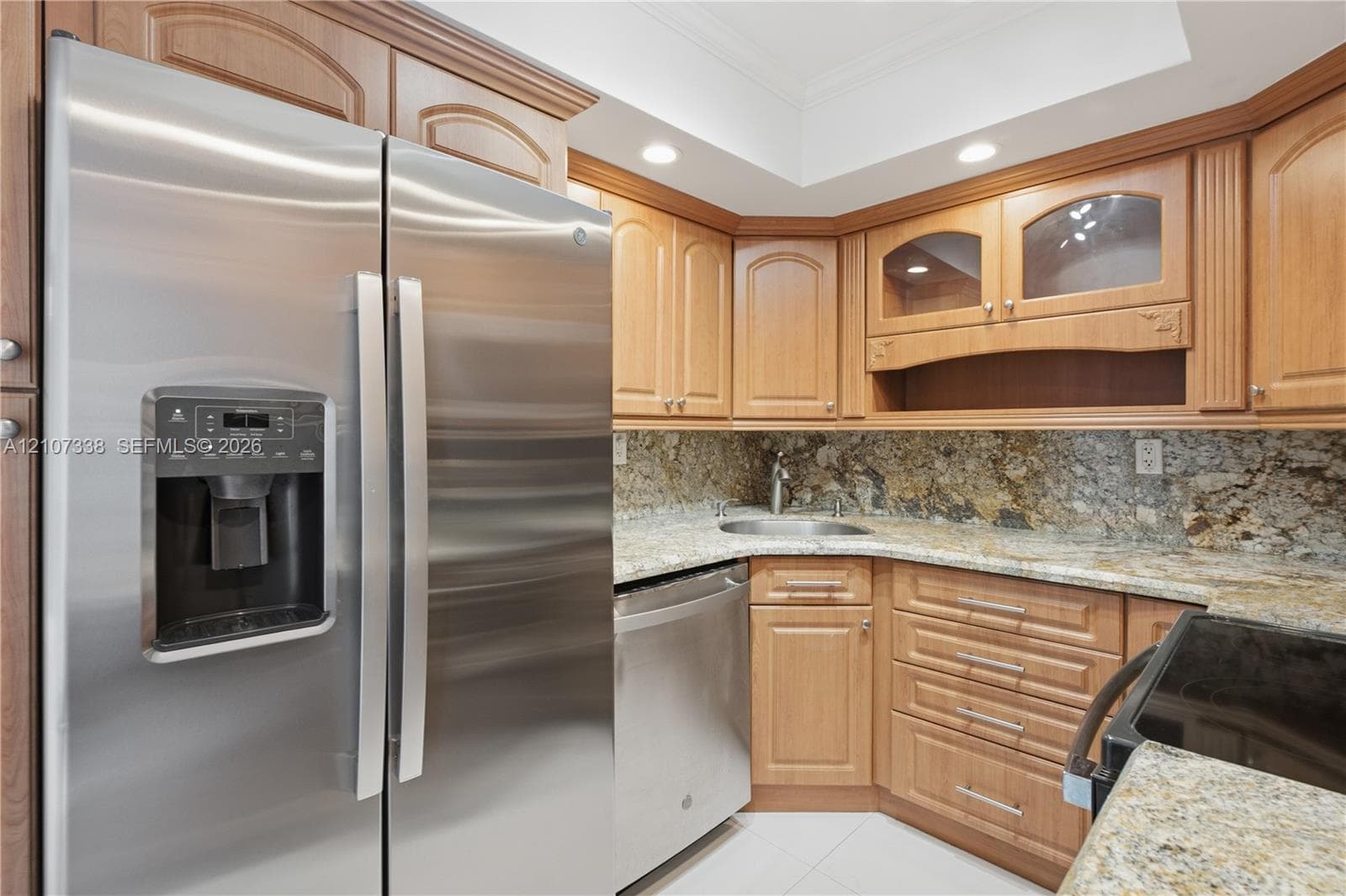 Stainless steel side-by-side refrigerator, natural stone countertops, tile backsplash, light wood-tone cabinetry with glass-front display accents, and recessed ceiling lighting