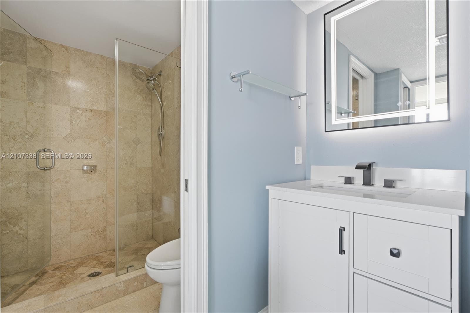 Bathroom featuring a white vanity with a quartz countertop, rectangular under-mount sink, matte black widespread faucet, and illuminated vanity mirror, alongside a walk-in shower with stone-tile surround and a glass door