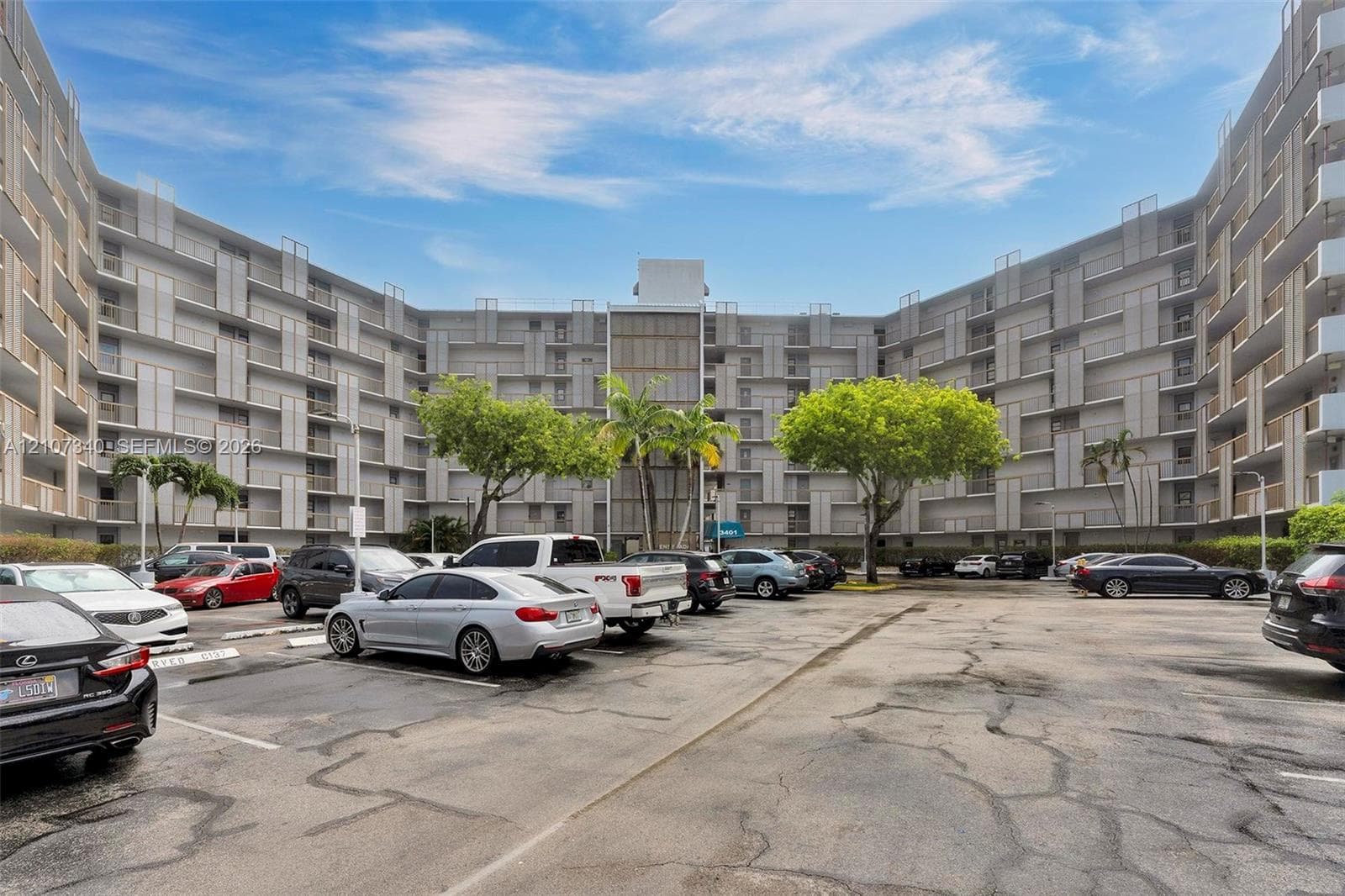 Multi-story residential building featuring a U-shaped architectural design, balcony enclosures, surface parking, and mature palm trees