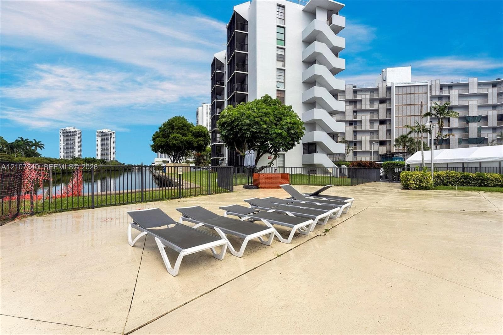 Concrete pool deck featuring a metal perimeter fence, water frontage, and mature landscaping