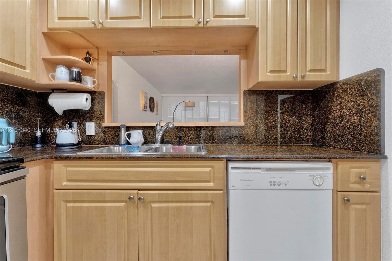 Kitchen featuring natural-finish wood cabinetry, stone-surface countertops, matching stone backsplash, dual-basin stainless steel sink with metal faucet, and built-in dishwasher