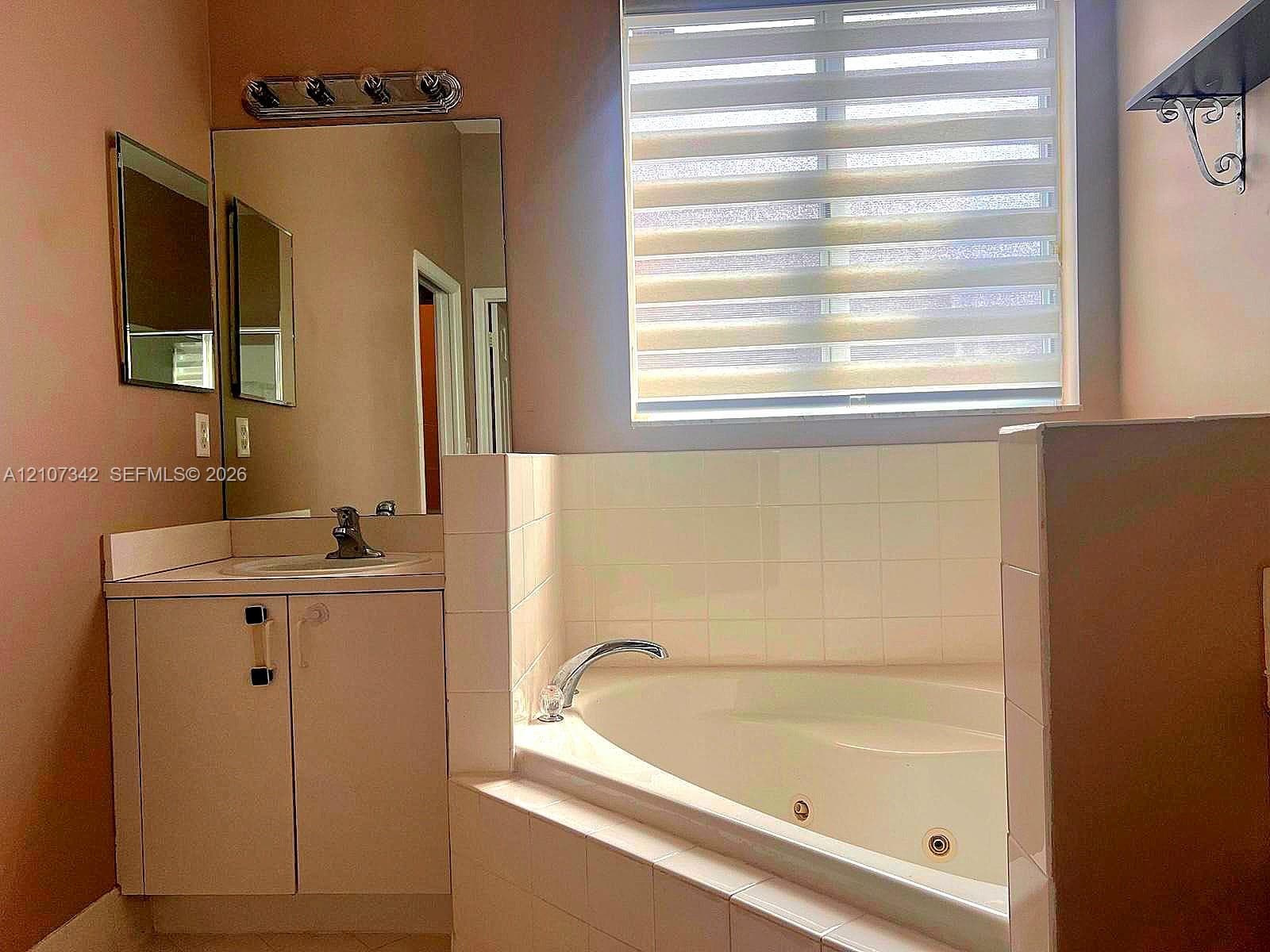 Bathroom featuring a soaking jetted tub with tile surround, vanity with inset sink, wall-mounted mirror, and tiered window shade