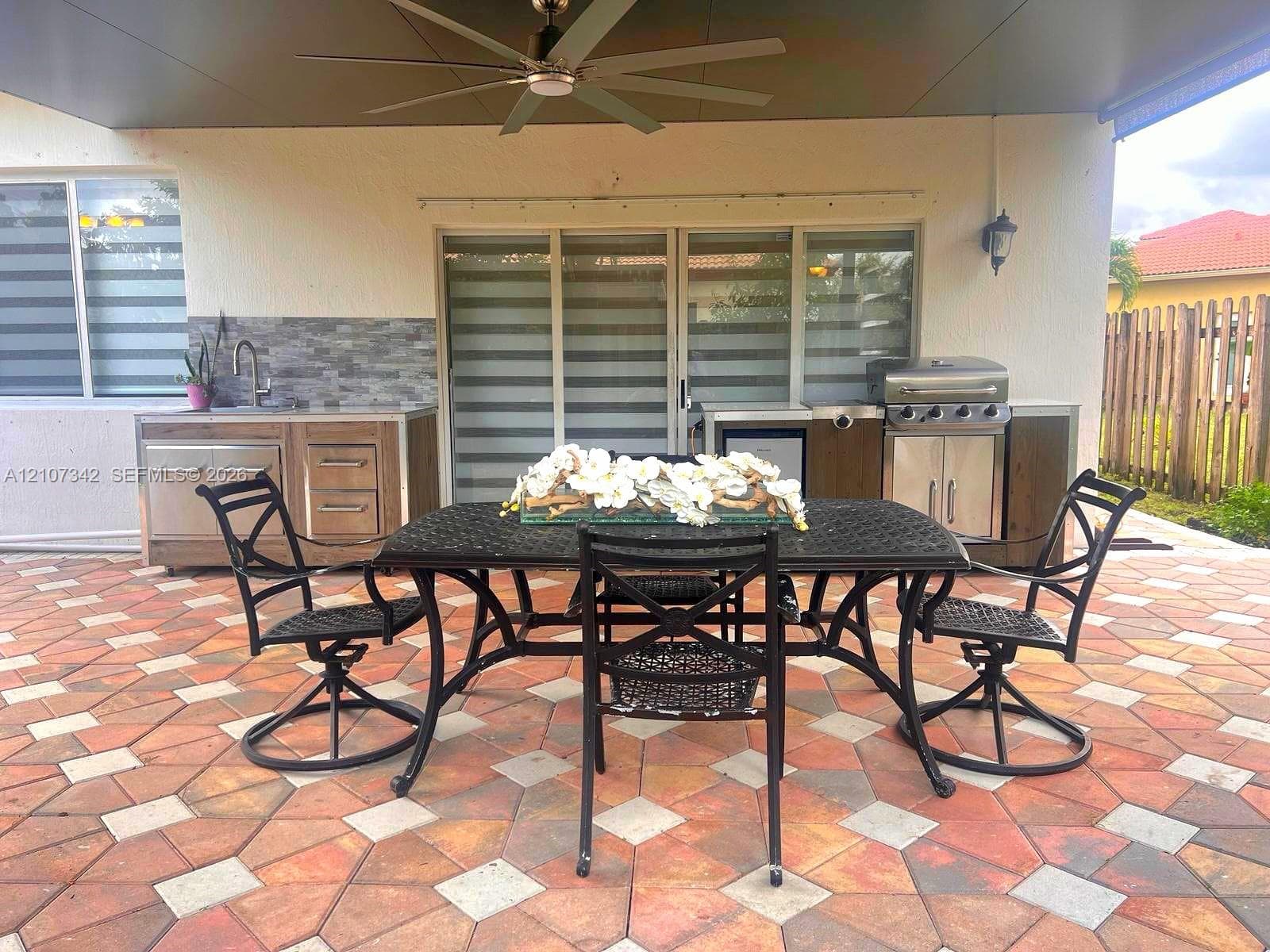 Covered patio featuring patterned stone-paver flooring, a stainless steel outdoor kitchen with a built-in grill and sink, stone-tile backsplash, and a ceiling-mounted fan