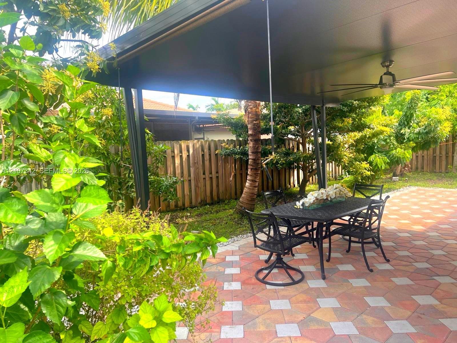 Covered patio featuring a multi-toned geometric paver floor, metal support columns, a mounted ceiling fan, and wooden perimeter fencing