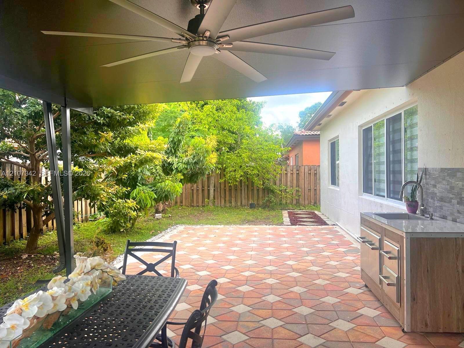 Covered patio featuring a ceiling fan, geometric paver flooring, built-in outdoor cabinetry with a sink, and a tiled backsplash