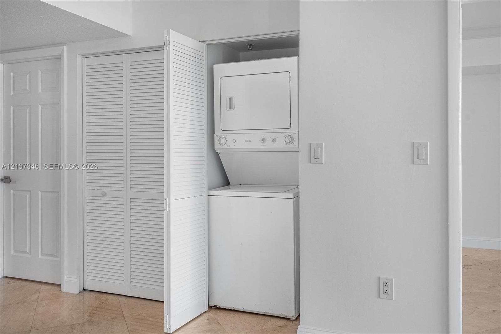 Stacked laundry unit housed within a closet featuring white louvered doors, offset by neutral-toned tiled flooring and a multi-panel door