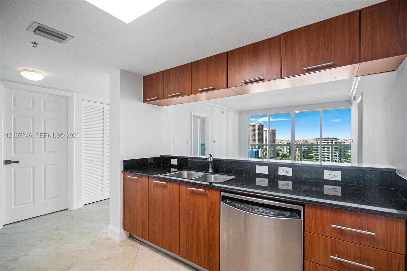Kitchen featuring wood-finish cabinetry, dark stone-surfaced countertops, stainless steel dishwasher, dual-basin stainless steel sink, and large windows with urban scenery