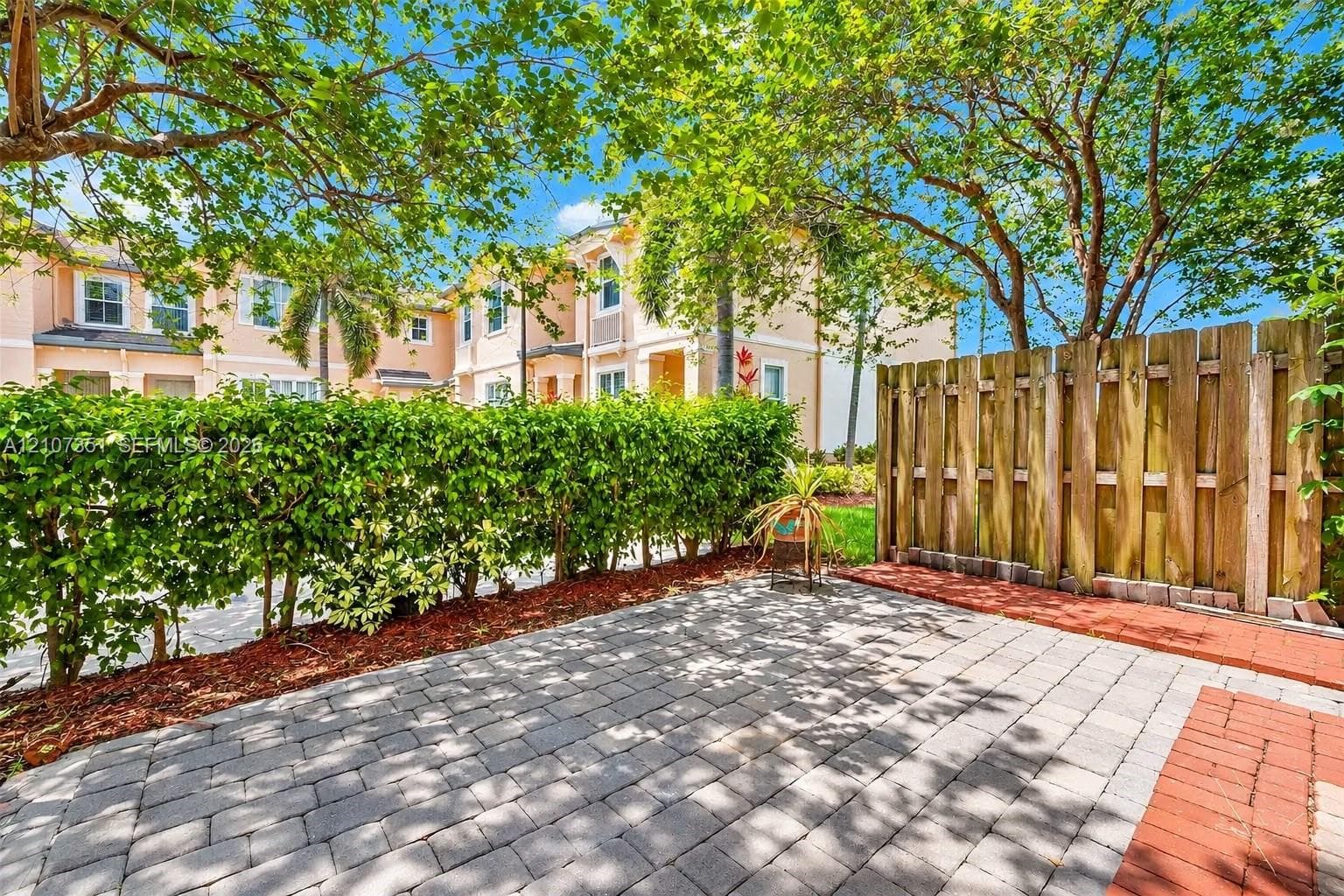 Exterior patio featuring interlocking concrete pavers, red brick border, privacy wood fencing, and perimeter hedging
