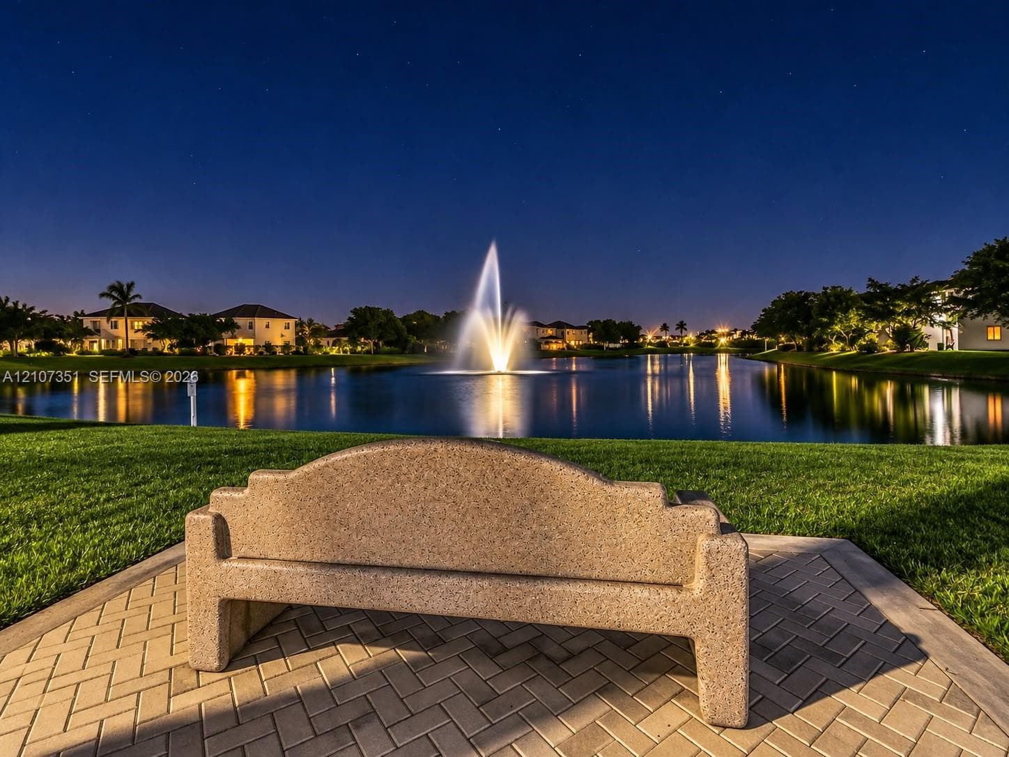 Concrete bench situated on a herringbone-pattern paver patio, overlooking a lake with a central water fountain and surrounding manicured turf