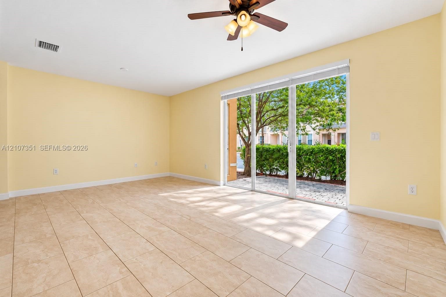 Spacious room featuring neutral tile flooring, a multi-blade ceiling fan with integrated light fixtures, and a glass sliding door opening to a paved patio area