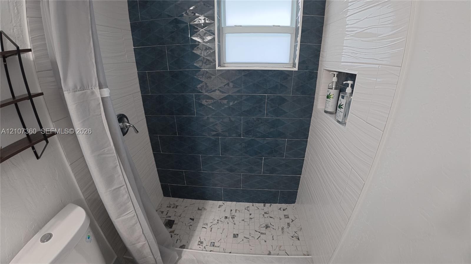 Shower area featuring blue geometric-patterned wall tiles, white textured wall tiles, a built-in recessed niche, and mosaic floor tile