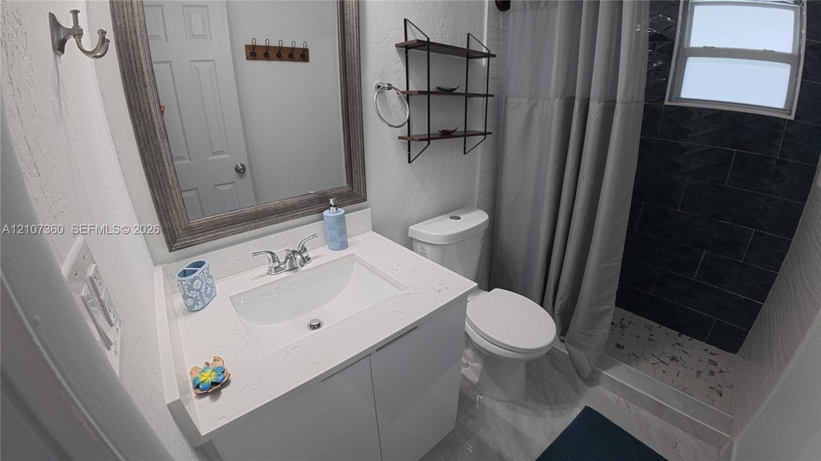 Bathroom featuring a vanity with a white composite countertop, a framed wall mirror, a shower with dark charcoal tile surround, and built-in metal shelving