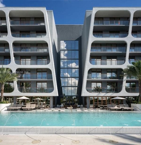 Contemporary architectural exterior featuring modular concrete framing, full-height glass curtain wall, integrated tiered balconies with glass railings, and a ground-level swimming pool with built-in water fountains